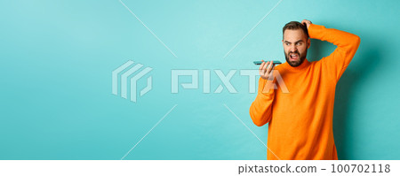 Confused man scratching head while talking on speakerphone, record voice message with indecisive face, standing in orange sweater over light blue background 100702118