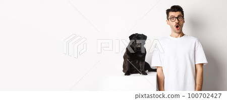 Image of dog owner and cute black pug looking at camera surprised and amazed, standing over white background 100702427