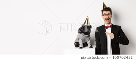 Happy dog owner and black pug wearing birthday party cones, man pointing at pug, standing over white background Happy dog owner and black pug wearing birthday party cones, man pointing at pug, standing over white background 100702451