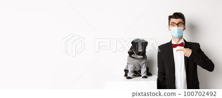 Coronavirus, pets and celebration concept. Amazed young man in face mask and suit pointing at cute black dog sitting near owner in party outfit, white background 100702492