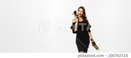 Beauty and fashion concept. Full length of attractive young woman using high heels like mobile phone, standing in black dress against white background 100702528