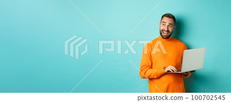 Handsome man doing online shopping, looking up thoughtful while using laptop, standing over light blue background Handsome man doing online shopping, looking up thoughtful while using laptop, standing over light blue background 100702545