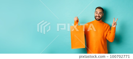 Happy young man showing peace sign and orange shopping bag, smiling pleased, standing over turquoise background Happy young man showing peace sign and orange shopping bag, smiling pleased, standing over turquoise background 100702771
