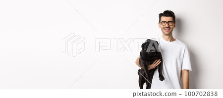 Handsome hipster guy holding his funny black pug dog, smiling at camera, standing over white background Handsome hipster guy holding his funny black pug dog, smiling at camera, standing over white background 100702838