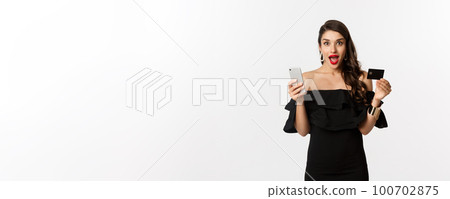 Online shopping concept. Fashionable woman in black dress, holding credit card with smartphone, looking excited, standing over white background 100702875