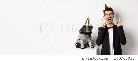 Cute black pug dog wearing party cone and standing near happy owner, man pointing fingers up at copy space, celebrating birthday, white background Cute black pug dog wearing party cone and standing near happy owner, man pointing fingers up at copy space, celebrating birthday, white background 100703033