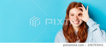 Headshot of cheerful redhead female model showing okay sign over eye, smiling satisfied and happy, standing against blue background 100703041