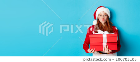 Happy holidays and Christmas concept. Cute redhead girl holding presents and pucker lips for kiss, wearing santa hat and funny sweater, blue background Happy holidays and Christmas concept. Cute redhead girl holding presents and pucker lips for kiss, wearing santa hat and funny sweater, blue background 100703103