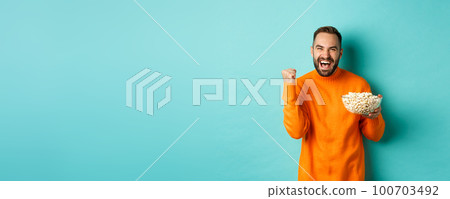 Cheerful handsome man in orange sweater, saying yes, cheering and rejoicing, eating popcorn and watching sports, making fist pump satisfied, standing over blue background Cheerful handsome man in orange sweater, saying yes, cheering and rejoicing, eating popcorn and watching sports, making fist pump satisfied, standing over blue background 100703492