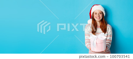 Winter and celebration concept. Beautiful redhead girl in santa hat wishing merry christmas, giving gift and smiling, standing over blue background Winter and celebration concept. Beautiful redhead girl in santa hat wishing merry christmas, giving gift and smiling, standing over blue background 100703541