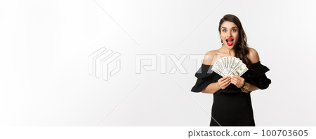 Beauty and shopping concept. Excited woman in black dress, showing money prize and staring happy at camera, standing over white background 100703605
