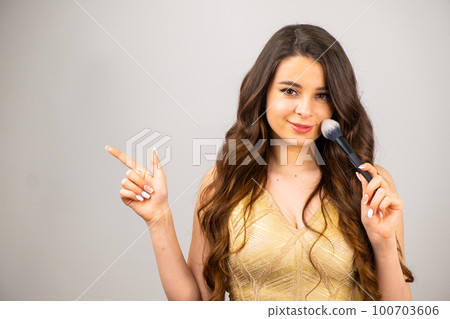 An attractive young woman holding makeup blusher brush and showing by finger to the copy space. An attractive young woman holding makeup blusher brush and showing by finger to the copy space. 100703606