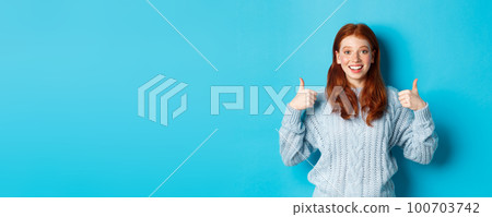 Cheerful teen girl with red hair, showing thumbs up in approval, like and praise gesture, standing over blue background 100703742