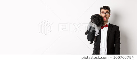 Funny young man in party suit, showing tongue and holding cute black pug on shoulder, celebrating with pet, standing over white background Funny young man in party suit, showing tongue and holding cute black pug on shoulder, celebrating with pet, standing over white background 100703794