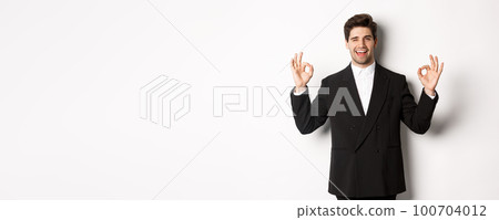 Concept of new year party, celebration and lifestyle. Portrait of successful handsome businessman in suit, winking and showing okay signs, recommending something good, white background Concept of new year party, celebration and lifestyle. Portrait of successful handsome businessman in suit, winking and showing okay signs, recommending something good, white background 100704012