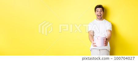 Concept of holidays and celebration. Happy man giving a gift and looking excited, standing against yellow background 100704027