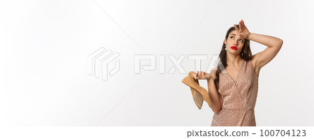 Party and celebration concept. Tired woman in elegant dress take off pair of heels, holding hand on forehead, express fatigue, standing over white background 100704123