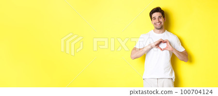 Happy romantic man showing heart sign, smiling and express love, standing over yellow background 100704124