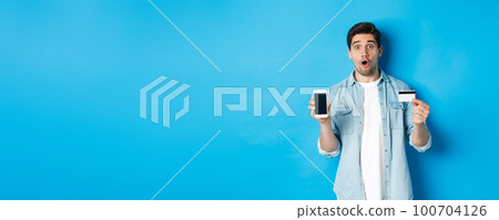 Amazed young man showing mobile cell phone screen and credit card, shop online, standing against blue background 100704126