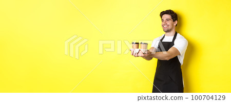 Friendly barista in black apron giving takeaway order, holding two cups of coffee and smiling, standing over yellow background Friendly barista in black apron giving takeaway order, holding two cups of coffee and smiling, standing over yellow background 100704129