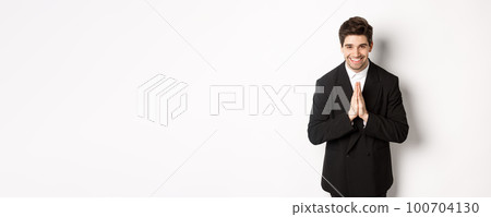 Portrait of handsome man in black suit, being grateful, saying thank you and bowing politely, smiling as holding hands together, expressing gratitude, standing over white background Portrait of handsome man in black suit, being grateful, saying thank you and bowing politely, smiling as holding hands together, expressing gratitude, standing over white background 100704130
