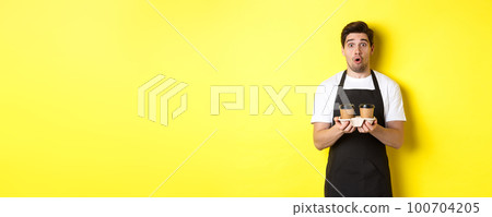 Barista serving coffee, looking surprised at camera, wearing black apron, standing against yellow background Barista serving coffee, looking surprised at camera, wearing black apron, standing against yellow background 100704205