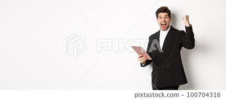 Portrait of handsome successful businessman in black suit, achieve goal, holding digital tablet and saying yes, standing against white background and rejoicing 100704316