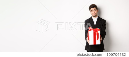 Concept of christmas holidays, celebration and lifestyle. Image of handsome boyfriend in black suit, giving you a present, extending hands with gift, standing over white background 100704382