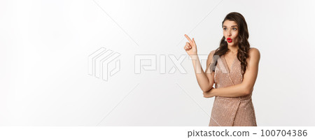 Christmas, holidays and celebration concept. Surprised girlfriend in evening dress, saying wow and pointing finger at promo offer, starring at logo, standing over white background 100704386