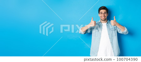 Attractive young man wearing glasses and casual clothes, showing thumbs up in approval, like something, standing against blue background 100704390