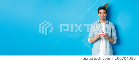 Smiling man holding b-day cake and wearing birthday party hat, celebrating over blue background 100704396