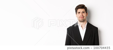Close-up of confused handsome man in suit, grimacing and looking perplexed, wearing trendy suit, standing against white background 100704435