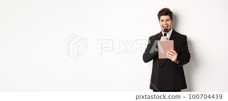Image of handsome man in black suit, singing karaoke on digital tablet, holding microphone, standing over white background 100704439