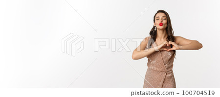 Celebration and party concept. Beautiful young woman in stylish dress, red lips, showing heart sign and waiting for kiss, standing over white background 100704519