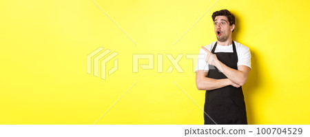 Waiter in black apron checking out promo offer, pointing finger and looking left at your logo, standing over yellow background 100704529