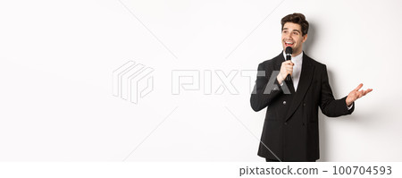 Portrait of handsome man in black suit singing a song, holding microphone and giving speech, standing against white background 100704593