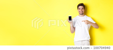 Image of attractive young man pointing finger at smartphone screen, showing an app, standing against yellow background 100704940