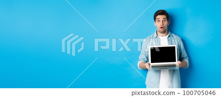 Man showing advertisement on laptop screen and looking amazed, saying wow and looking at camera, standing against blue background 100705046