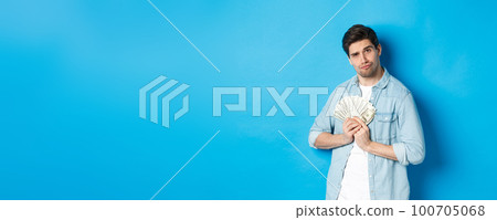 Handsome young man keeping money to himself, smiling and looking greedy, standing over blue background Handsome young man keeping money to himself, smiling and looking greedy, standing over blue background 100705068