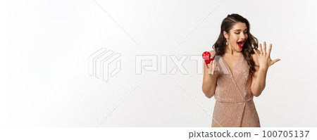 Excited young woman in glamour dress being engaged, showing finger with ring and looking happy, receive marriage proposal, standing over white background 100705137