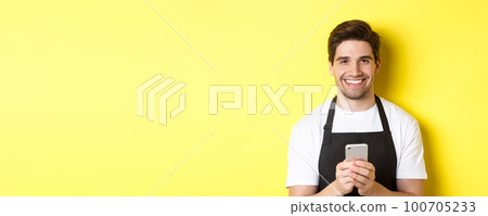 Close-up of handsome barista sending message on mobile phone, smiling happy, standing over yellow background Close-up of handsome barista sending message on mobile phone, smiling happy, standing over yellow background 100705233