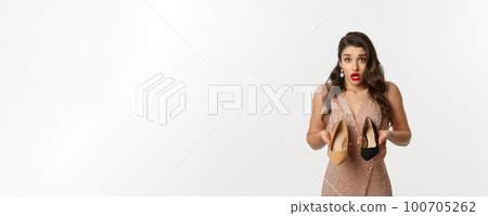 Party and celebration concept. Beautiful woman in elegant dress asking help, showing two different heels, clueless what to wear, standing over white background 100705262