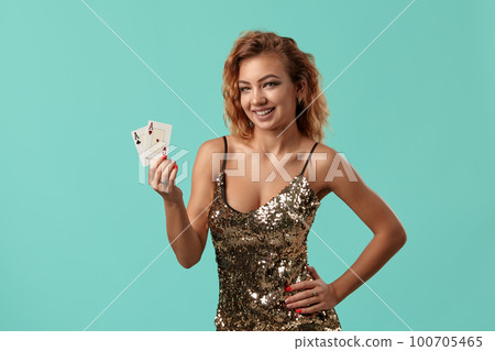 Ginger girl in shiny golden dress is posing holding two playing cards in her hands standing against a blue studio background. Casino, poker. Close-up. Ginger girl in shiny golden dress is posing holding two playing cards in her hands standing against a blue studio background. Casino, poker. Close-up. 100705465