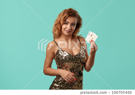 Ginger girl in shiny golden dress is posing holding two playing cards in her hands standing against a blue studio background. Casino, poker. Close-up. Ginger girl in shiny golden dress is posing holding two playing cards in her hands standing against a blue studio background. Casino, poker. Close-up. 100705480