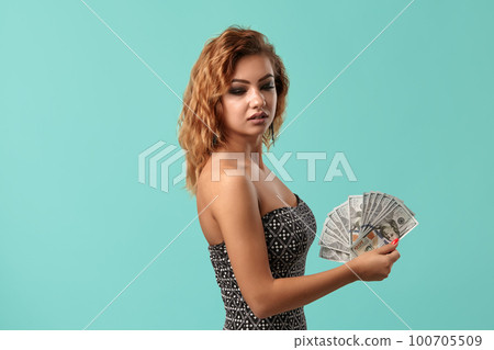 Ginger girl wearing gray dress is posing holding some cash in her hands standing against a blue studio background. Casino, poker. Close-up. Ginger girl wearing gray dress is posing holding some cash in her hands standing against a blue studio background. Casino, poker. Close-up. 100705509