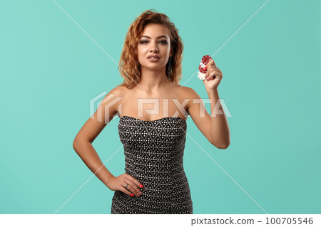 Ginger girl wearing gray dress is posing holding some chips in her hands standing against a blue studio background. Casino, poker. Close-up. 100705546