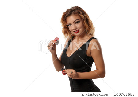 Ginger girl wearing a black dress is posing holding red and green chips in her hands standing isolated on white background. Casino, poker. Close-up. 100705693