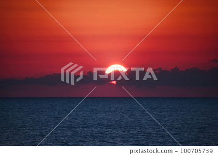 Dramatic sunset over the ocean horizon at dusk with clouds partly covering the sun against a deep red sky background - dreamy glowing light fading into the waters Dramatic sunset over the ocean horizon at dusk with clouds partly covering the sun against a deep red sky background - dreamy glowing light fading into the waters 100705739