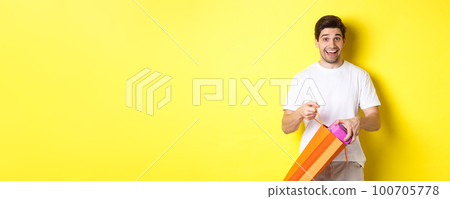 Concept of holidays and celebration. Happy and surprised man take out gift and looking at camera, standing over yellow background 100705778