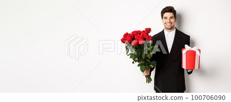 Concept of holidays, relationship and celebration. Handsome boyfriend in black suit, holding bouquet of red roses and a gift, wishing merry christmas, standing over white background 100706090